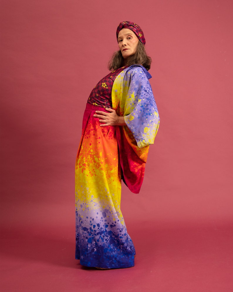 K&ouml;nnte beinhalten: Eine Person tr&auml;gt ein farbenfrohes Outfit mit einem Regenbogen-Ombre-Muster. Das Outfit besteht aus einem lang&auml;rmeligen Oberteil mit Sternenmuster, einer Hose und einem Turban. Die Person steht vor einem rosa Hintergrund.