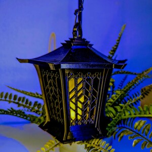 May include: A black metal lantern with a geometric design hangs from a chain. The lantern has a yellow light inside, casting a warm glow. The lantern is surrounded by green ferns.