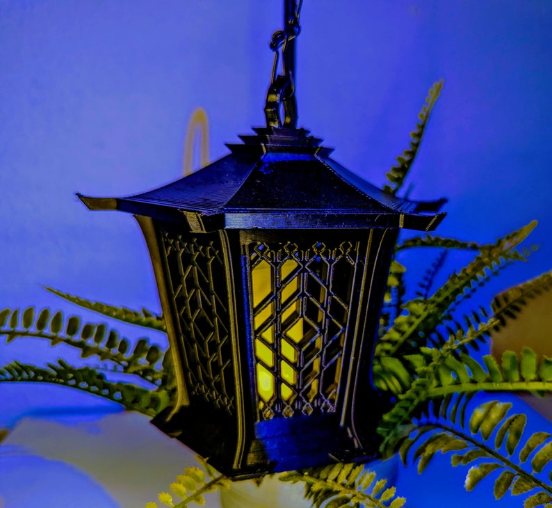 May include: A black metal lantern with a geometric design hangs from a chain. The lantern has a yellow light inside, casting a warm glow. The lantern is surrounded by green ferns.