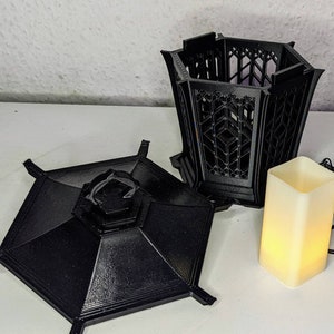 May include: A black, 3D printed, hexagonal lantern with a removable lid and a decorative, geometric design. The lantern is sitting on a white surface next to a small, white, battery-operated candle.