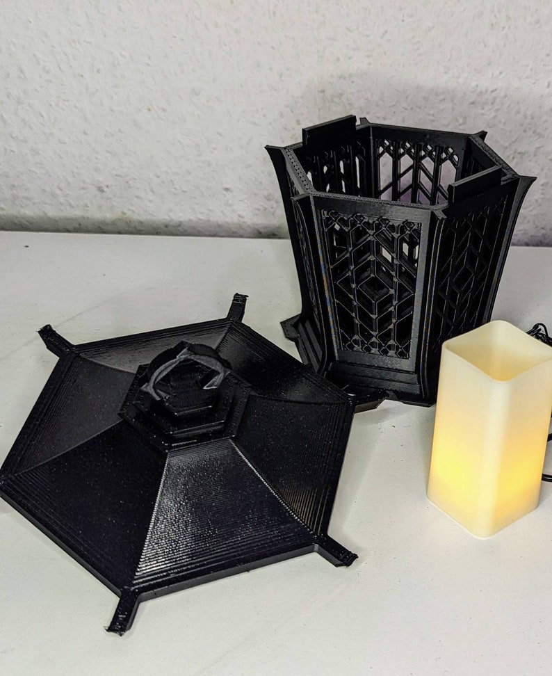 May include: A black, 3D printed, hexagonal lantern with a removable lid and a decorative, geometric design. The lantern is sitting on a white surface next to a small, white, battery-operated candle.