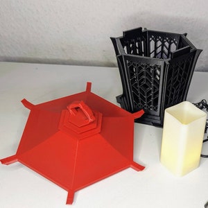 May include: A red hexagonal lantern with a black lattice design. The lantern is made of plastic and has a handle on the top. The lantern is sitting on a white surface.