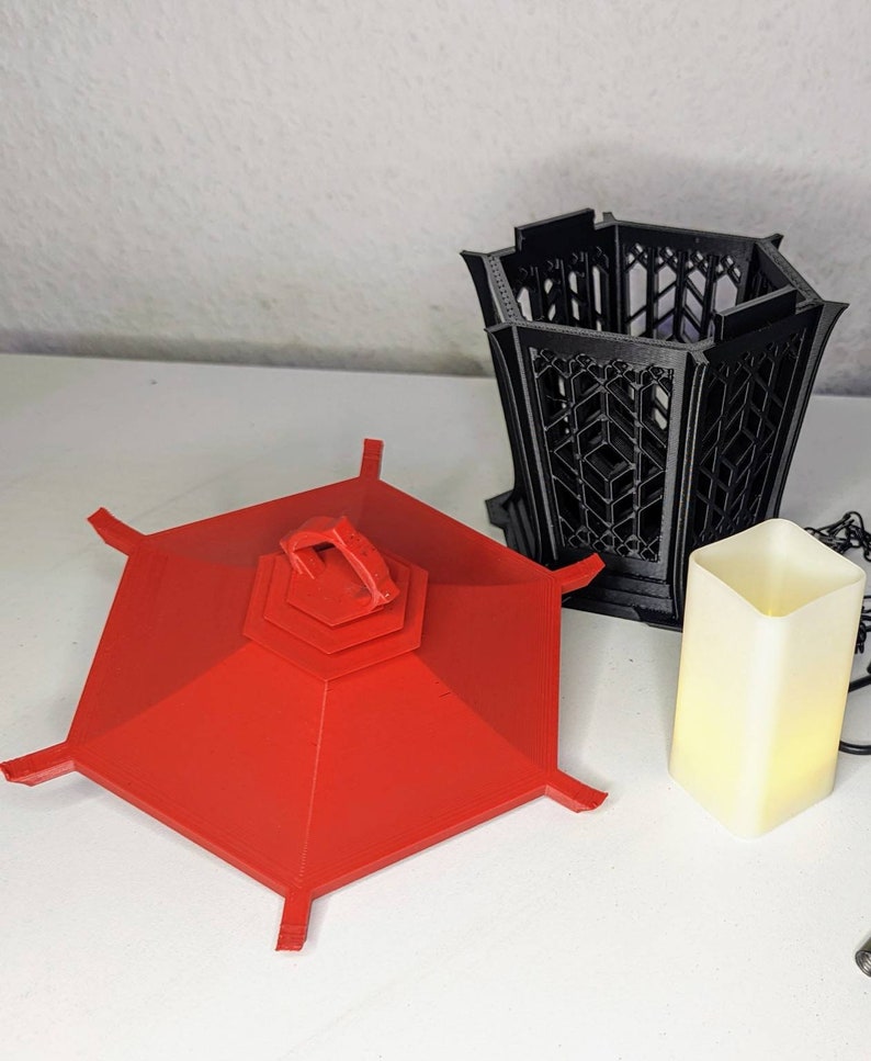 May include: A red hexagonal lantern with a black lattice design. The lantern is made of plastic and has a handle on the top. The lantern is sitting on a white surface.