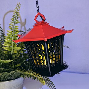 May include: A black and red hanging lantern with a geometric design. The lantern has a red roof and a black body with a lattice pattern. The lantern is lit with a warm yellow light.