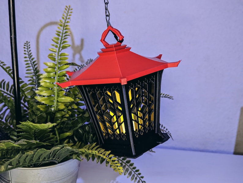 May include: A black and red hanging lantern with a geometric design. The lantern has a red roof and a black body with a lattice pattern. The lantern is lit with a warm yellow light.
