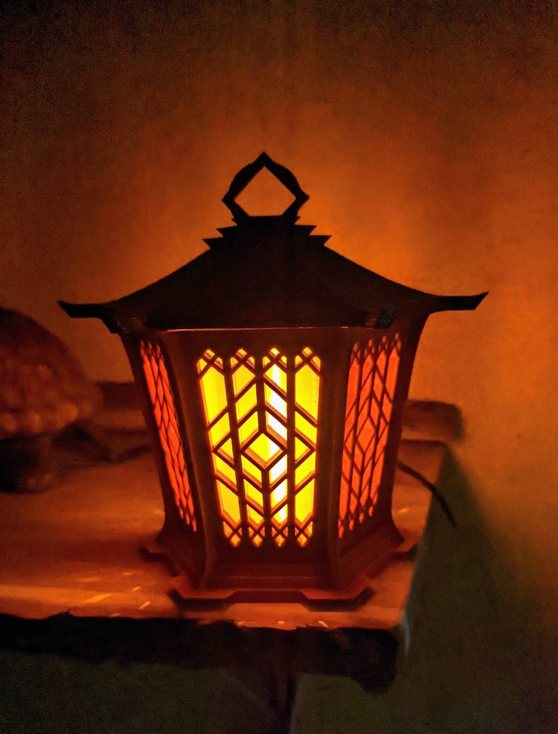May include: A decorative lantern with a black metal frame and a warm orange glow emanating from the inside. The lantern has a geometric design with intricate cutouts.