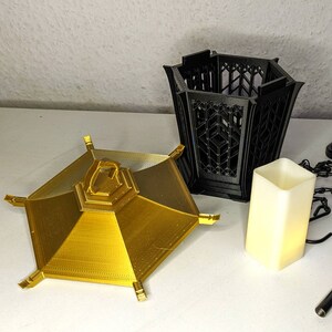 May include: Two 3D printed candle holders. One is gold and has a hexagonal shape with a raised center. The other is black and has a hexagonal shape with an open top.