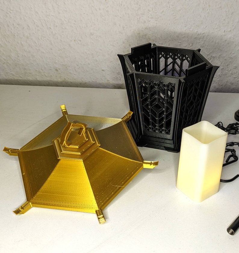 May include: Two 3D printed candle holders. One is gold and has a hexagonal shape with a raised center. The other is black and has a hexagonal shape with an open top.