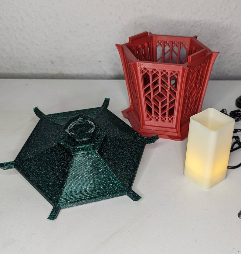 May include: A set of three 3D printed decorative pieces. The largest piece is a red hexagonal lantern with a lattice design. The second piece is a green hexagonal top with a textured surface. The smallest piece is a white rectangular candle holder.
