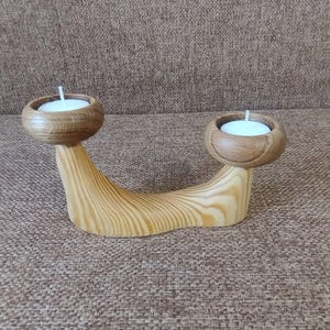 Peut inclure: Un bougeoir double en bois clair avec une base incurvée lisse présentant des motifs de grain prononcés. Deux coupelles circulaires en bois de ton plus foncé sont placées à chaque extrémité, contenant chacune une petite bougie chauffe-plat.