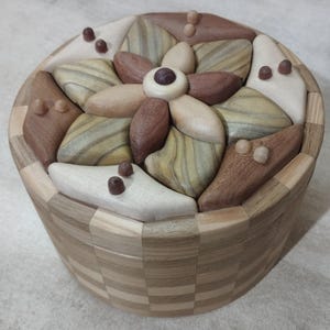 Könnte beinhalten: Eine runde Holzschatulle mit einem Deckel, der mit einem Intarsien-Blumenmuster aus verschiedenen hellen, mittleren und dunklen Holztönen verziert ist. Die Seiten der Schatulle sind in einem Schachbrettmuster aus abwechselnden hellen und dunklen Holzsegmenten gefertigt.