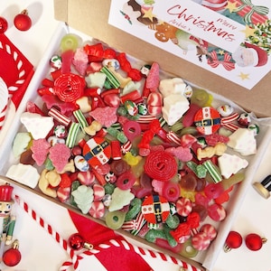 Puede incluir: Una caja rebosante de una colorida variedad de caramelos con temática navideña. Los caramelos incluyen dulces rojos, verdes y blancos, incluyendo formas de goma, caramelos duros y golosinas envueltas. Una tarjeta festiva con "Merry Christmas" es visible.