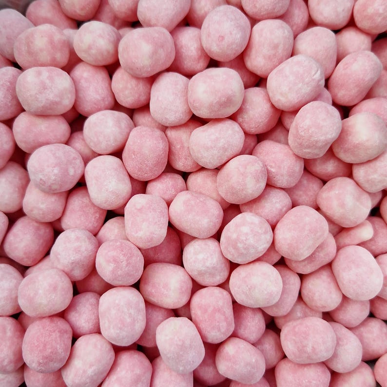May include: A close-up shot of a pile of pink candy. The candies are small, oval-shaped, and have a slightly rough texture. The color is a soft, pastel pink with some white patches, suggesting a sugary coating. The candies are tightly packed together.