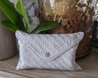 May include: A white and silver patterned fabric pouch with a button closure. The pouch is made of a soft, textured material.