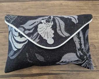 May include: A rectangular envelope-style pouch featuring a black and grey floral jacquard pattern. The flap is edged with white piping and secured by a single grey snap button closure. The fabric has a textured weave throughout the design.