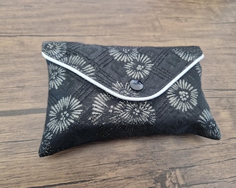 May include: A rectangular black fabric pouch featuring a silver floral pattern and geometric quilted lines. The envelope-style flap is secured with a single black button and finished with a thin white piped edge along the closure.