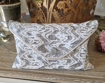 May include: A rectangular envelope-style pouch featuring a textured white and silver metallic jacquard fabric with an abstract wavy pattern. The flap is secured with a single centred grey button and finished with a thin white piping trim.