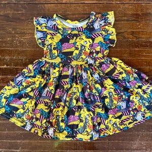 Puede incluir: Un vestido amarillo con un estampado negro, turquesa, rosa y morado. El vestido presenta un cuello redondo, mangas cortas con volantes y una falda acampanada. El estampado incluye personajes y elementos de una película de animación popular.