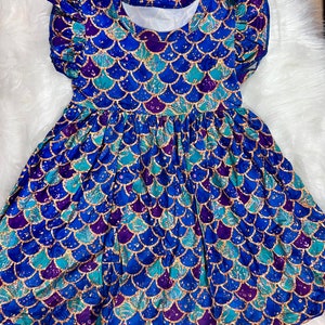 Könnte beinhalten: Ein blaues und türkisfarbenes Kleid mit Meerjungfrauen-Schuppenmuster und goldenen Glitzerakzenten. Das Kleid hat kurze, gekräuselte Ärmel und einen gerafften Rock.