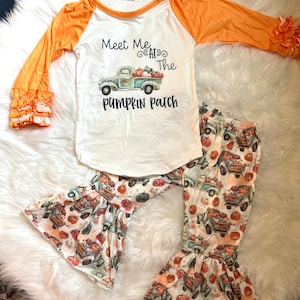 以下が含まれることがあります： かぼちゃ畑のデザインが施された白とオレンジのロングスリーブシャツ。シャツには「Meet Me at the Pumpkin Patch」の文字と、かぼちゃを積んだビンテージトラックが描かれています。シャツは、かぼちゃ畑のプリントが施された白いパンツと組み合わせています。