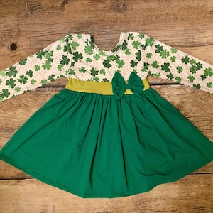 May include: A green and white dress with a shamrock print and a green bow. The dress has long sleeves and a green skirt.