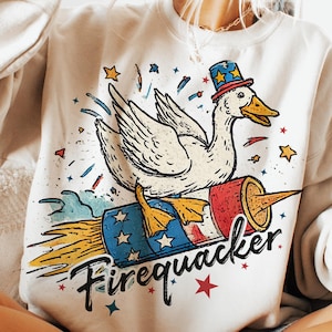 May include: Cream-colored sweatshirt featuring a cartoon duck riding a rocket. The duck wears a patriotic hat and the rocket has red, white, and blue accents. The text "Firequacker" is below the duck.