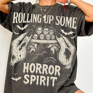 Puede incluir: Camiseta gris oscuro con el texto "ROLLING UP SOME HORROR SPIRIT". El gráfico presenta un par de manos sosteniendo una colección de personajes clásicos de películas de terror. Murciélagos y una serpiente también están incluidos.