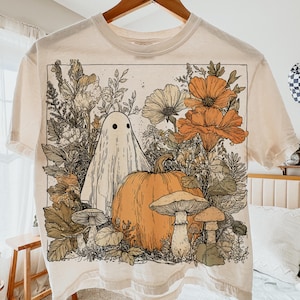 May include: Cream-colored t-shirt featuring a Halloween-themed graphic. The design includes a white ghost, an orange pumpkin, mushrooms, and flowers in shades of orange, green, and beige. The shirt is hanging on a wooden hanger.