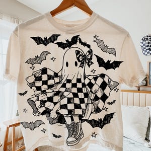 May include: Cream-coloured t-shirt with a black and white ghost design. The ghost wears a checkered cape and bow, surrounded by bats and stars. The shirt is hanging on a wooden hanger.