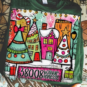Puede incluir: Sudadera verde con un gráfico colorido con temática navideña. El diseño presenta casas estilizadas, árboles de Navidad y el texto "38008 Bolivar, Tenesse". La obra de arte incluye un bastón de caramelo, estrellas y cajas de regalo.
