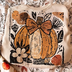 May include: Cream-coloured sweatshirt with an orange pumpkin design and a bow, surrounded by black leaves and flowers. The pumpkin has a textured, linocut style. The design is centered on the front of the sweatshirt, evoking an autumnal theme.