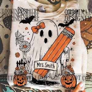 May include: Beige Halloween-themed sweatshirt with a ghost design holding a large orange pencil. The ghost has a bow and the name "Mrs. Smith" on a banner. Includes pumpkins, flowers, bats, and spiderwebs.