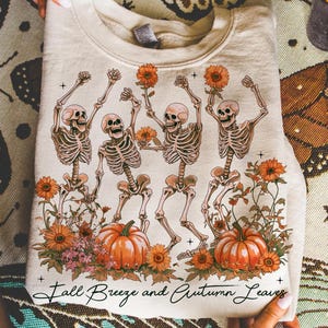 May include: Beige sweatshirt with a graphic of dancing skeletons holding sunflowers, pumpkins, and floral elements. The text "Fall Breeze and Autumn Leaves" is printed at the bottom. A seasonal design, perfect for autumn.