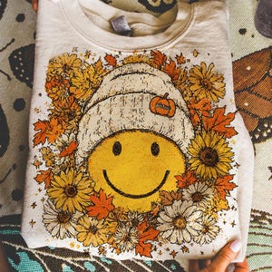 Puede incluir: Sudadera beige con una cara sonriente que lleva un gorro blanco y una calabaza. El diseño está rodeado de flores y hojas de otoño en tonos amarillos, naranjas y blancos. Un diseño con temática otoñal.