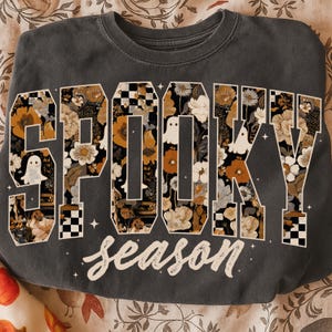 以下が含まれることがあります： 「SPOOKY SEASON」の文字が花柄でデザインされたダークグレーのスウェットシャツ。文字はオレンジ、白、黒の花で埋められ、小さなゴーストのイラストが描かれています。「season」の文字は筆記体で書かれています。