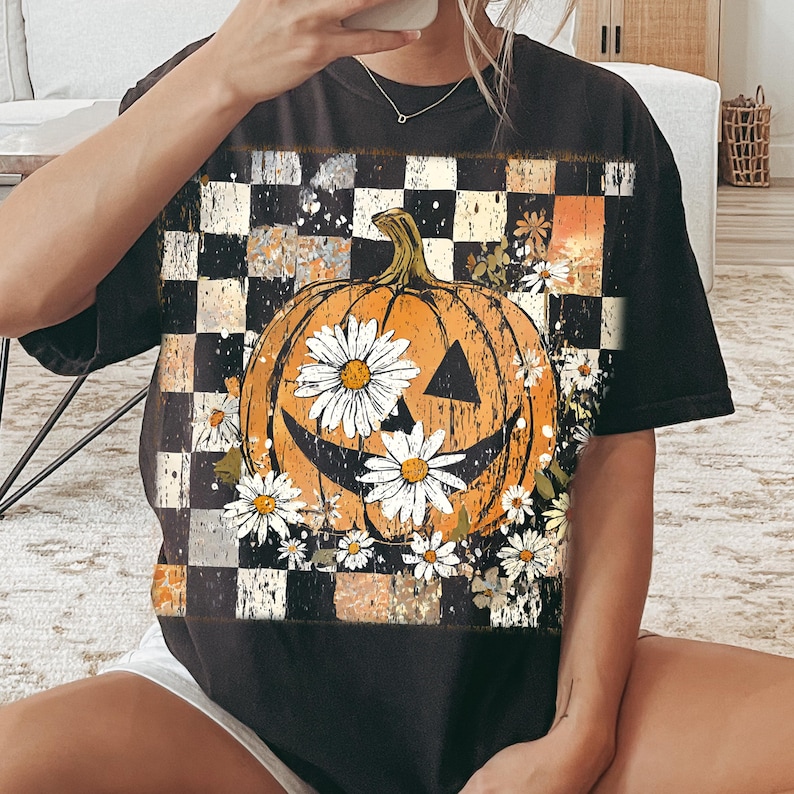 May include: Black t-shirt featuring a smiling orange pumpkin with daisy flowers and a checkered background. The pumpkin has triangle eyes and a curved mouth. The daisies are white with yellow centers. The shirt is a relaxed fit.
