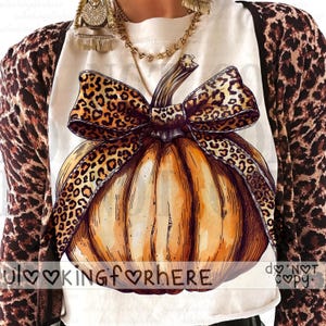 May include: A white t-shirt featuring a large orange pumpkin graphic with a leopard print bow. The shirt is worn under a leopard print cardigan and a gold chain necklace. The text "LOOKINGfORHERE" and "DO NOT COPY" is printed across the image.