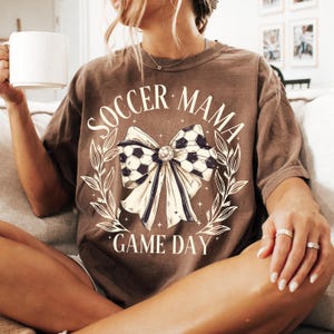 Puede incluir: Camiseta marr&oacute;n con un lazo de bal&oacute;n de f&uacute;tbol blanco y el texto "Soccer Mama Game Day" impreso en ella.
