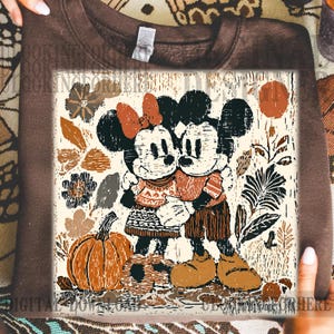 May include: A brown sweatshirt with a graphic print of Mickey and Minnie Mouse hugging, set against a backdrop of autumn leaves and a pumpkin. The design uses brown, orange, and cream tones, evoking a warm, seasonal feel. The sweatshirt is a digital download.