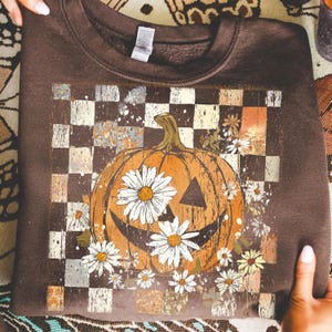May include: A brown sweatshirt featuring a smiling jack-o'-lantern design with white daisy flowers. The pumpkin is set against a checkered background with a distressed, vintage look. A perfect fall or Halloween themed apparel.