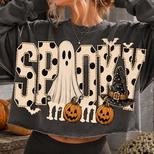 Può includere: Una felpa grigio antracite a maniche lunghe con orlo grezzo che presenta la scritta SPOOKY MOM in grandi lettere beige a pois neri. Il design include un fantasma bianco, due zucche arancioni, un cappello da strega e dei piccoli pipistrelli.