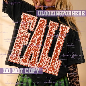 May include: A black short-sleeved t-shirt featuring the word FALL in large, distressed, burnt orange block letters with a white speckled pattern. The design is centered on the chest, and the shirt is worn over a long-sleeved leopard print top.