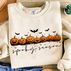 Op de afbeelding: Cr&egrave;mekleurige sweatshirt met een rij uitgesneden oranje pompoenen en zwarte vleermuissilhouetten. De tekst "spooky season" staat in zwart schrift eronder, perfect voor Halloween.