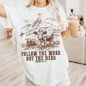 May include: White t-shirt with a brown graphic of a cow and two calves in a field with mountains in the background. The graphic also includes a cross on a mountaintop. The text "FOLLOW THE WORD NOT THE HERD" is below the graphic, with "ISAIAH 8:11-13" below the text.