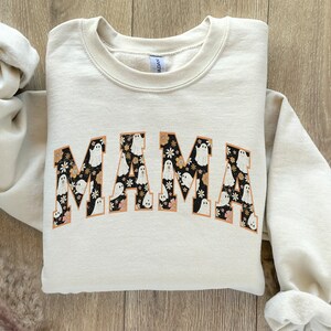 Pu&ograve; includere: Felpa color crema con la scritta "MAMA" in un font retr&ograve;. Le lettere sono riempite di fantasmi bianchi e motivi floreali su sfondo nero, bordati di arancione.