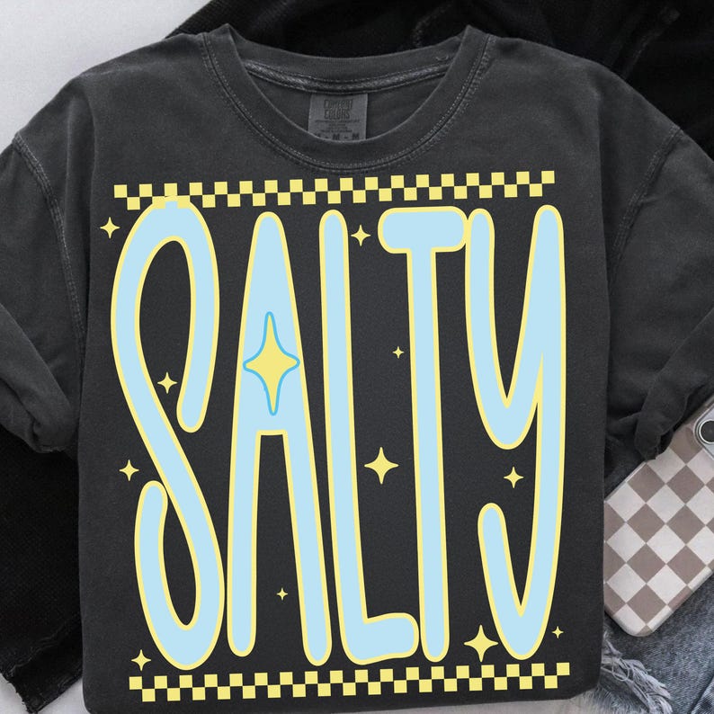 Puede incluir: Camiseta gris oscuro con la palabra "SALTY" en azul claro, delineada en amarillo. La palabra tiene una estrella en el centro y est&aacute; enmarcada por un patr&oacute;n de cuadros amarillos y blancos. Peque&ntilde;as estrellas amarillas est&aacute;n dispersas alrededor de la palabra.