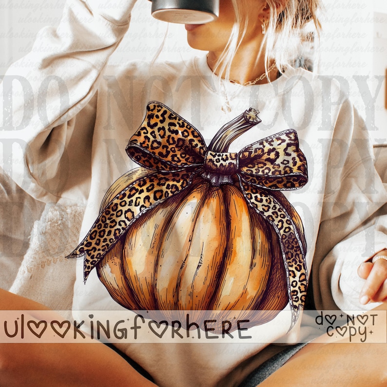 May include: A cream-colored crewneck sweatshirt featuring a large graphic illustration of an orange pumpkin adorned with a prominent leopard print bow. The pumpkin design includes detailed black line work and shading, centered on the front of the garment.