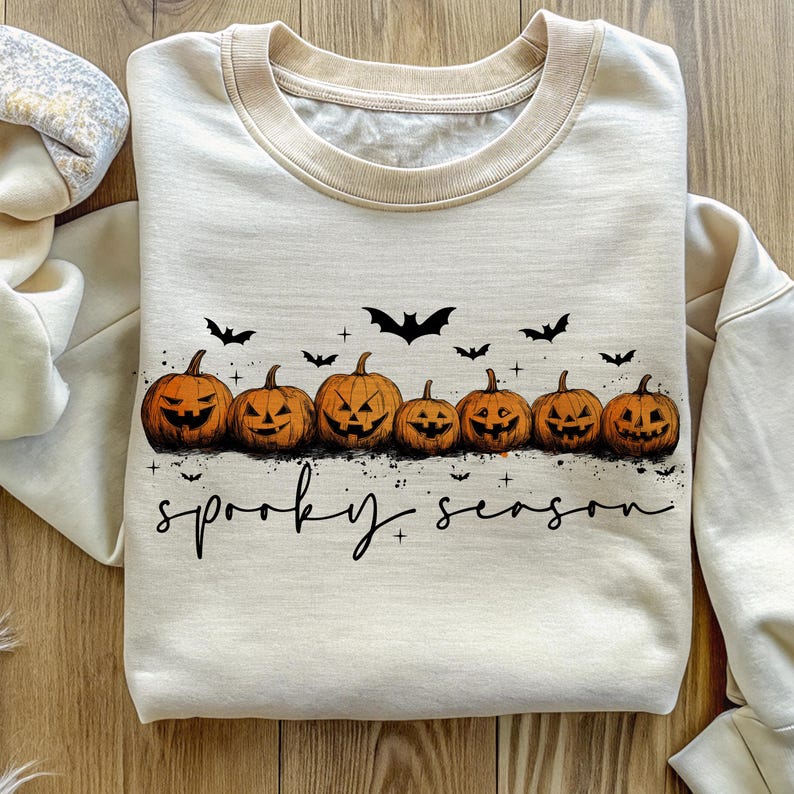 Op de afbeelding: Cr&egrave;mekleurige sweatshirt met een beige kraag en manchetten. De voorkant heeft een rij uitgesneden oranje pompoenen met zwarte vleermuizen erboven en de tekst "spooky season" in een scriptlettertype.