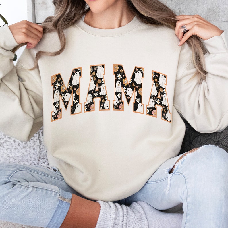 Pu&ograve; includere: Felpa color crema con la scritta "MAMA" in un font in stile collegiale. Le lettere sono riempite con un motivo floreale nero e illustrazioni di fantasmi bianchi, delineate in marrone. Un design festivo per Halloween.