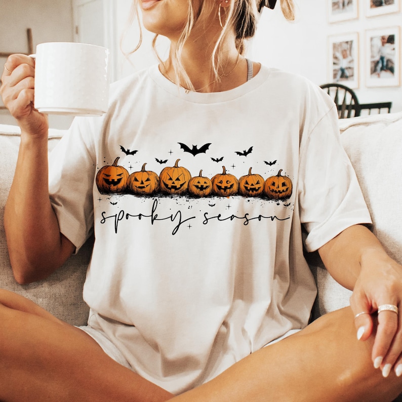 Op de afbeelding: Cr&egrave;mekleurig T-shirt met een rij uitgesneden oranje pompoenen met zwarte gezichten en vliegende vleermuizen erboven. De woorden "spooky season" staan in zwarte letters onder de pompoenen. Een witte mok wordt in de linkerhand gehouden.